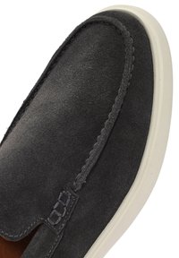 Zapato gris de ante tipo slip-on con suela de goma blanca en contraste. Presenta costuras visibles y una textura suave. Diseño limpio y minimalista.