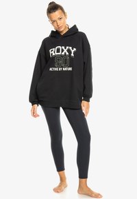 Schwarzer Kapuzenpullover mit dem Grafikdruck "ROXY 90 ACTIVE BY NATURE", lockere Passform und langen Ärmeln, kombiniert mit dunklen Leggings.