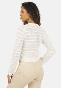 Biały sweter dzianinowy z teksturowanym, prążkowanym wzorem; ma okrągły dekolt i prążkowany dół. Zestawiony z jasnymi, dopasowanymi spodniami.