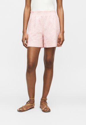 Femme portant un short rose clair brodé et des sandales plates à brides couleur cuir clair, debout sur un fond blanc.