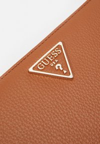 Braunes Lederportemonnaie mit einem goldenen, dreieckigen Logo und dem Text "GUESS USA" sowie einem Fragezeichen. Texturierte Oberfläche mit sichtbarem Maserungsmuster.