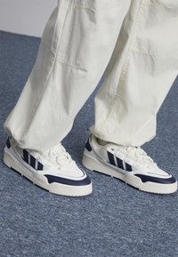 Vita och marinblå sneakers med texturerat läder, som har tre marinblå ränder på sidan och en tjock vit sula.