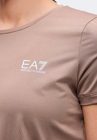 Κοντινό πλάνο μιας φανέλας με κοντά μανίκια χρώματος ταουπ, με λευκό λογότυπο EA7 Emporio Armani στο πάνω μέρος του στήθους.