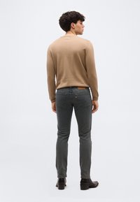 Άνδρας στέκεται με την πλάτη γυρισμένη, φορώντας ανοιχτό καφέ πουλόβερ, γκρι στενά τζιν και μαύρες μπότες με τακούνι, σε απλό λευκό φόντο.