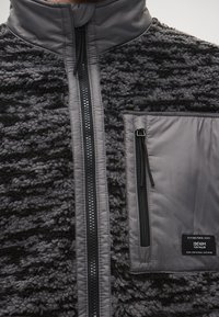 Fleecová bunda so zmiešanou šedou textúrou, s predným zipsom, vysokým golierom a náprsné vrecko s logom. Mäkký, teplý materiál.