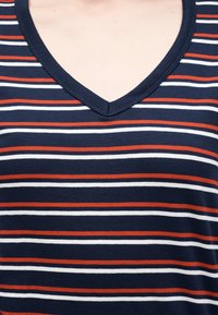 Κοντινό πλάνο ενός μπλουζιού σε ναυτικό μπλε με λαιμόκοψη V και οριζόντιες λευκές και σκουροπορτοκαλί ρίγες στο πάνω μέρος του στήθους και του λαιμού ενός ατόμου.
