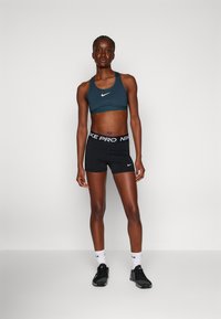Reggiseno sportivo di un verde scuro con il logo Nike bianco, abbinato a pantaloni corti neri con un bordo "Nike Pro" bianco. Scarpe da ginnastica nere e calze bianche.