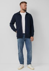Marineblauwe zip-up jas met een staande kraag, ribgebreide manchetten en voorzakken, gecombineerd met lichtblauwe jeans en beige sneakers.