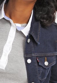 Chaqueta de mezclilla con herrajes plateados, color azul oscuro y detalles de costura. Lleva encima una camisa gris con cuello blanco y detalles en azul.