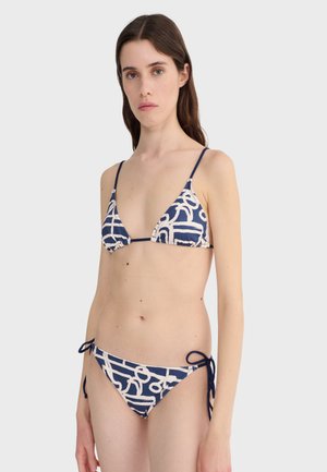 Femme portant un bikini string à motifs abstraits bleu marine et blanc avec des bonnets triangles et un bas à nouer sur les côtés, debout devant un fond uni.
