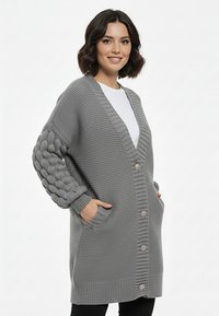 Szary sweter na drutach z dekoltem w serek, teksturowanym ciałem, rękawami z splotem warkoczowym i dużymi guzikami. Posiada dwie przednie kieszenie.