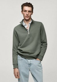 Felpa olive green con zip al collo, maniche lunghe, texture morbida e polsini a coste, abbinata a jeans in denim blu chiaro.