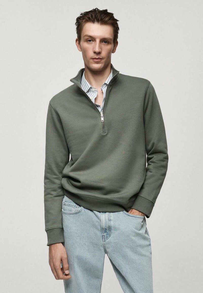 Felpa olive green con zip al collo, maniche lunghe, texture morbida e polsini a coste, abbinata a jeans in denim blu chiaro.