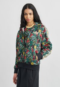 Frau trägt eine schwarze, mit Blumenmuster versehene Jacke mit Reißverschluss, gelben Streifen und schwarze Hose, steht vor einem schlichten weißen Hintergrund.