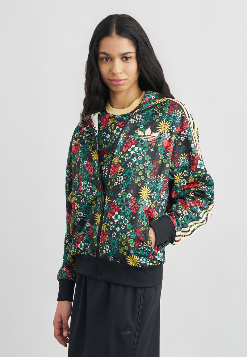 Frau trägt eine schwarze, mit Blumenmuster versehene Jacke mit Reißverschluss, gelben Streifen und schwarze Hose, steht vor einem schlichten weißen Hintergrund.