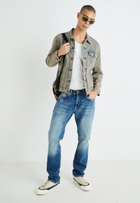Giacca di denim grigio sbiadito, t-shirt bianca, jeans azzurri chiari e sneakers color crema con accenti blu scuro. Gli accessori includono occhiali da sole gialli.