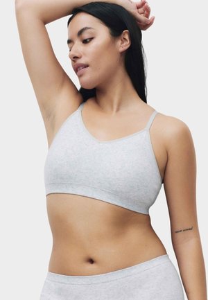 Vrouw draagt een lichtgrijze naadloze bralette en bijpassende broekjes, met één arm omhoog en kijkt naar beneden aan haar zijde.