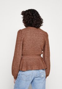 Femme aux cheveux bouclés foncés portant un pull en laine marron avec des épaules bouffantes et un jean bleu clair usé, vue de dos sur un fond blanc.