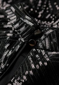 Tissu noir avec des rayures verticales en sequins argentés et deux boutons noirs brillants fermant le vêtement texturé.