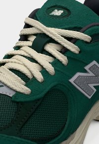 Πράσινο αθλητικό παπούτσι New Balance με κρεμ κορδόνια, υφή από δίχτυ και σουέντ, και μεγάλο αντανακλαστικό λογότυπο «N» στο πλάι.
