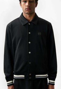 Giacca sportiva nera, tessuto a trama, bottoni a pressione, logo esagonale sul petto, polsini e orlo a righe a costine. Design del colletto semplice.