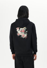 Felpa nera con cappuccio, caratterizzata da una grafica dettagliata di un teschio con rose e il testo "Santa Cruz" sul retro. Materiale in cotone.