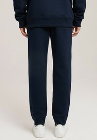 Granatowe dresy wykonane z miękkiego materiału, z elastycznymi mankietami i tylną kieszenią. Połączone z białymi trampkami, widok z tyłu.