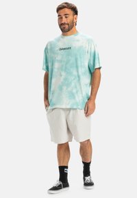Light blue tie-dye t-shirt with short sleeves and black "Quiksilver" logo, paired with beige shorts and black socks with white accents.