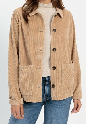 Femme portant une veste beige en velours côtelé boutonnée avec deux poches avant sur un col roulé beige clair et un jean bleu.