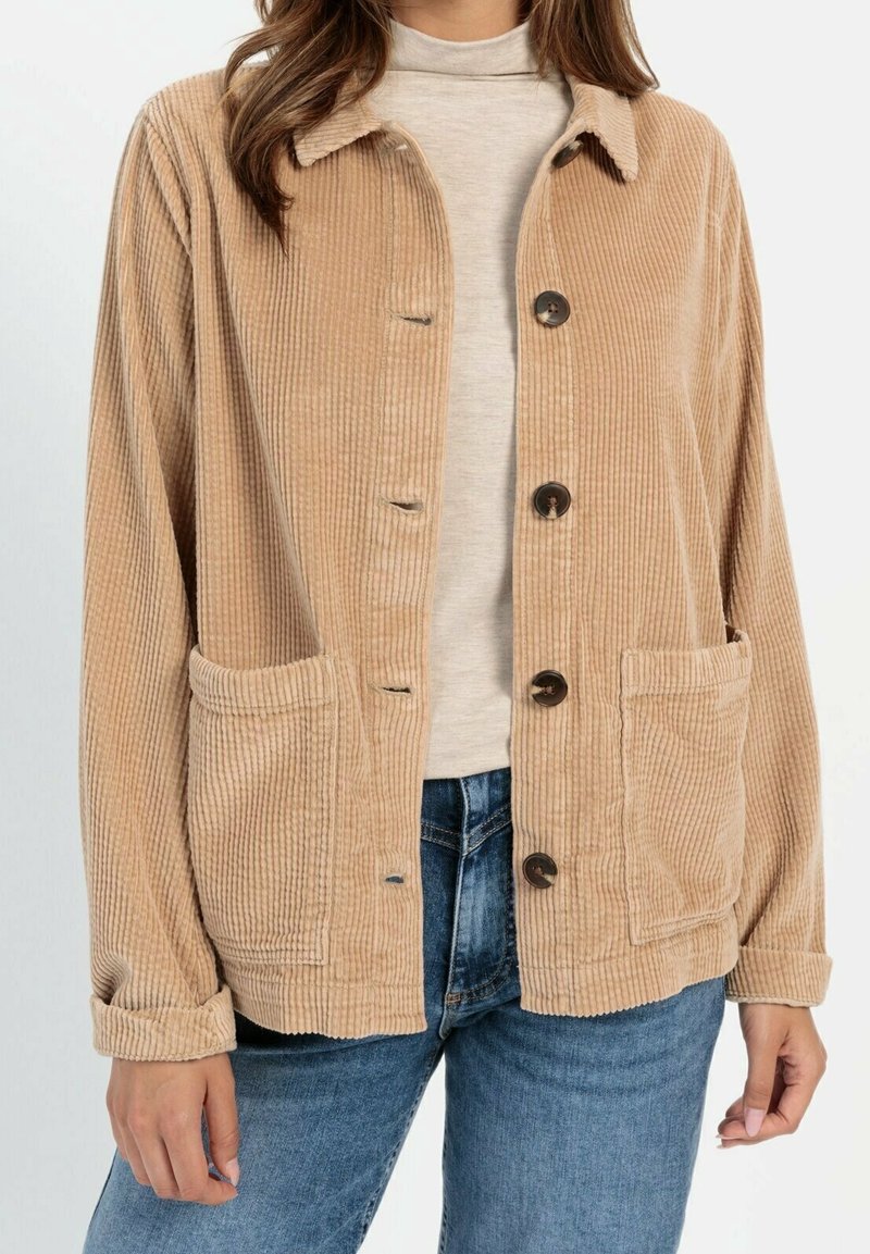 Femme portant une veste beige en velours côtelé boutonnée avec deux poches avant sur un col roulé beige clair et un jean bleu.