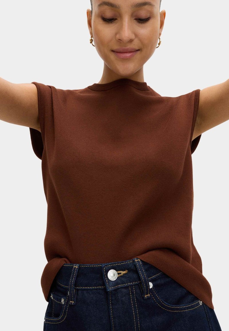 Jeune femme portant un haut marron sans manches et un jean bleu foncé, avec des boucles d'oreilles dorées en forme d'anneau, les bras levés, les yeux fermés et un léger sourire.