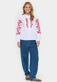 Hvid langærmet bluse med rødt blomsterbroderi på ærmerne, knaplukning, kombineret med løse blå denimjeans og flade sko med leopardmønster.