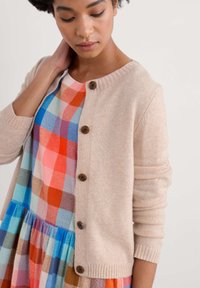 Helle beigefarbene Strickjacke mit Holzknöpfen, getragen über einem bunten karierten Kleid in Rottönen, Blautönen und Grüntönen. Strukturierten Stoff.