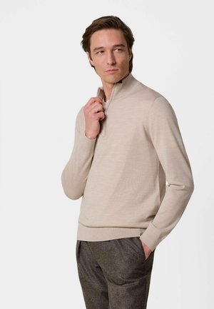 Homme portant un pull beige à fermeture éclair quart, pantalon gris, une main ajustant la fermeture éclair, l'autre dans la poche, regardant sur le côté.