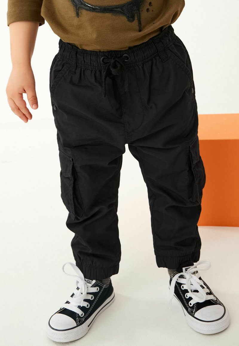 Enfant portant un pantalon cargo noir avec des poignets élastiques et des baskets noires avec des lacets blancs, debout devant un fond clair avec un bloc orange à proximité.