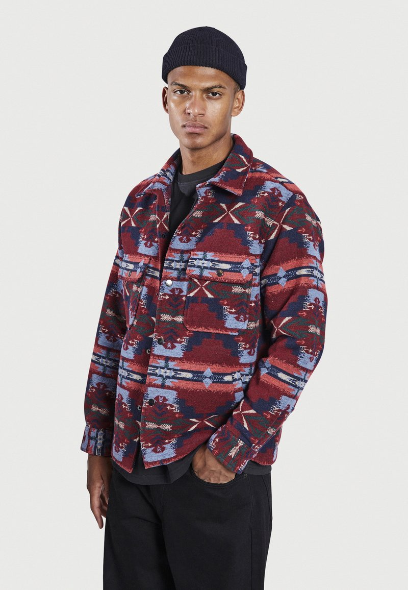 Veste en flanelle à motifs rouges et bleus avec boutons-pression, deux poches avant et un col classique ; tissu texturé avec des motifs géométriques.