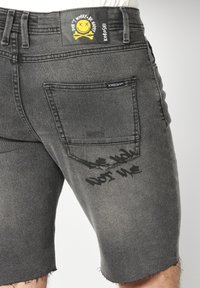 Short en denim gris foncé avec une poche arrière, une étiquette jaune représentant un visage souriant avec l'inscription "Don't Worry Be Happy" et un texte noir estompé près de la poche.