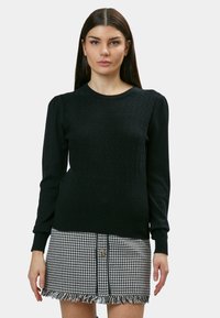 Schwarzer Strickpullover mit strukturiertem Zickzackmuster und langen Ärmeln, kombiniert mit einem schwarz-weißen Hahnentritt-Rock mit fransigem Saum.