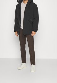 Svart isolerad jacka med huva, dragkedjeförsedda fickor och en avslappnad passform, kombinerad med bruna rutig byxor och ljusa sneakers.