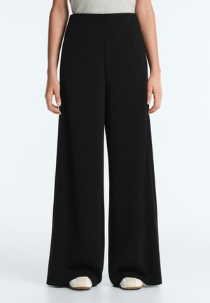 Persoană care poartă pantaloni negri cu picioare largi și pantofi albi, stând în fața unui fundal alb simplu, cu mâinile relaxate pe lângă corp.