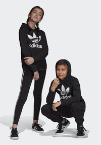 Mustad mustad Adidas kapuutsiga dressid ja retuusid, millel on valge logo ja triibud. Paari musti sneaks'e valgel taustal. Kaks last poseerivad koos.