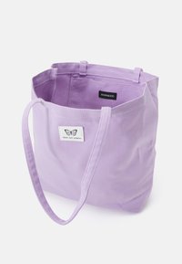 Sac fourre-tout en toile violet avec deux poignées. Présente une étiquette en tissu avec un logo de papillon et le texte "Pensez, ressentez, imaginez." Design simple.