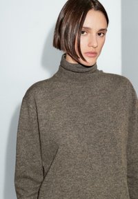 Frau mit glattem braunem Bob-Haarschnitt, die einen braunen Rollkragenpullover vor einer schlichten hellgrauen Wand trägt.