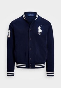 Navyblå varsityjacka med vita randiga detaljer, knappar och fickor. Har en stor vit logotyp på bröstet och en nummer "3" på ärmen.