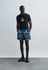 Black short-sleeve t-shirt with gold logo, paired with blue patterned shorts featuring white accents, and black sneakers. Smooth texture.