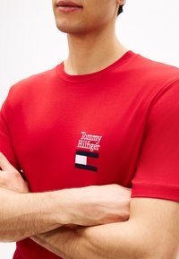 Άνδρας που φοράει κόκκινο μπλουζάκι Tommy Hilfiger με κεντημένο λογότυπο στο στήθος, με σταυρωμένα χέρια, μπροστά σε απλό λευκό φόντο.