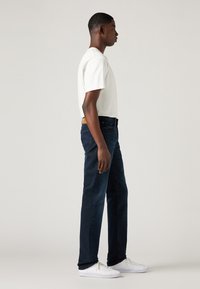 Fiatal férfi oldalról áll, fehér pólót, sötétkék farmert és fehér sportcipőt visel, egy egyszínű háttér előtt.