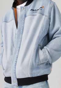 Jasnoniebieska bomberka z denimu z czarnym ściągaczem w pasie. Na lewej piersi znajduje się logo McLaren, a po bokach kieszenie.