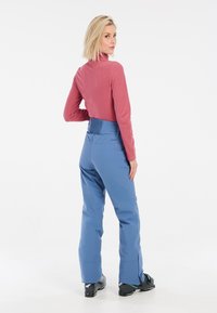 Light pink long-sleeve top paired with blue ski pants. Pants have a textured waistband and flared legs, with black ski boots visible.