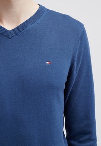 Niebieski sweter z dzianiny z dekoltem w serek. Posiada małe, wielokolorowe logo na lewej piersi. Miękka tekstura, dopasowane rękawy.